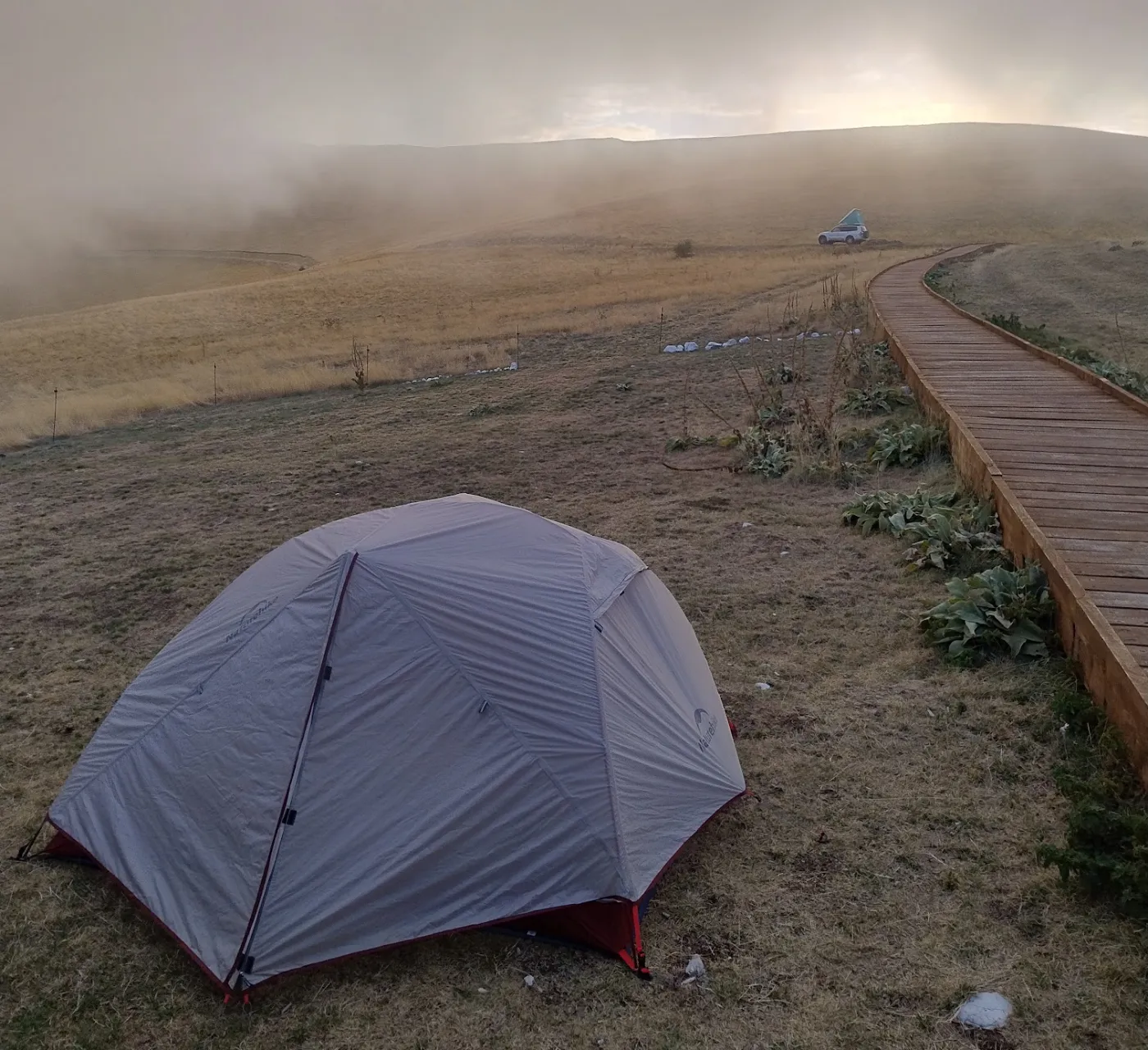 A grey dome tent on dry grass beside a wooden boardwalk that curves away into thick fog over the Sibillini mountains